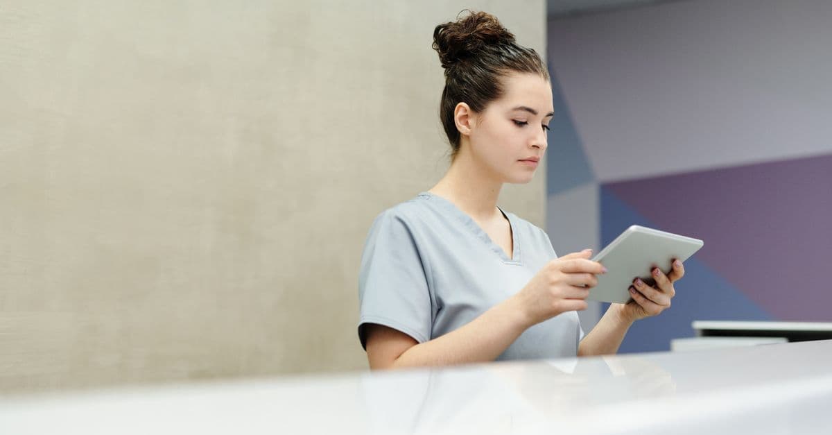 A receptionist in scrubs using a tablet at an office reception desk.